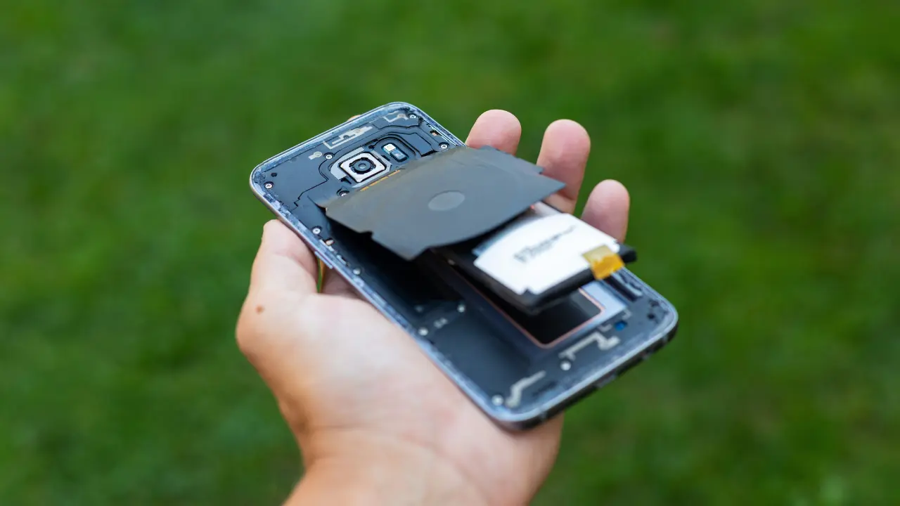 Close de uma bateria de celular estufada sendo examinada por um técnico em uma bancada de assistência técnica.